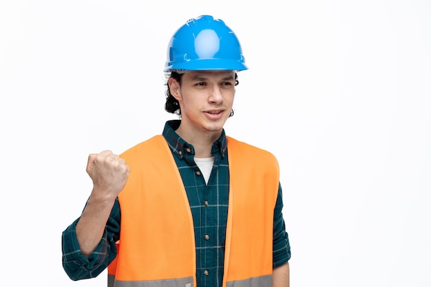 Giovane ingegnere maschio scontento che indossa casco di sicurezza e giubbotto di sicurezza guardando il lato tenendo il pugno in aria isolato su sfondo bianco