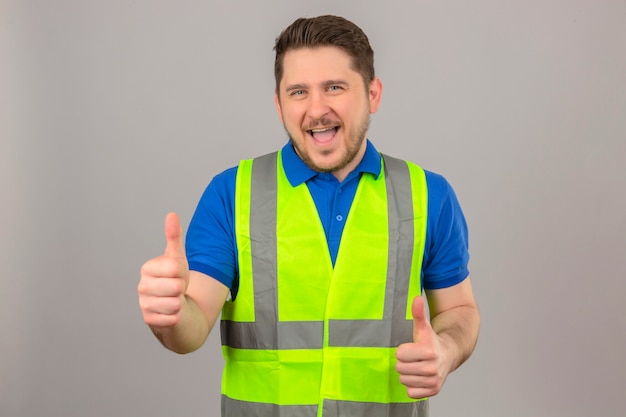 Giovane ingegnere uomo che indossa la maglia della costruzione che guarda l'obbiettivo con un grande sorriso e la faccia felice che mostra i pollici in su sopra fondo bianco isolato