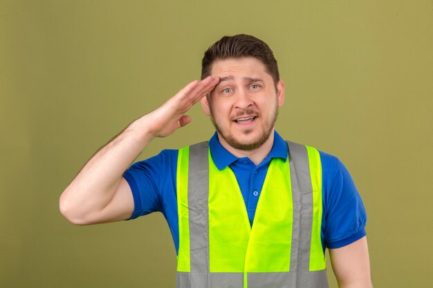 Giovane ingegnere uomo che indossa la maglia di costruzione guardando la fotocamera con un sorriso fiducioso salutando con la mano sopra la testa isolato su sfondo verde