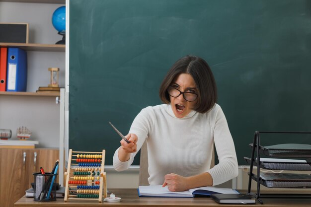 giovane insegnante arrabbiata che indossa occhiali punta alla telecamera con il puntatore seduto alla scrivania con gli strumenti della scuola in classe