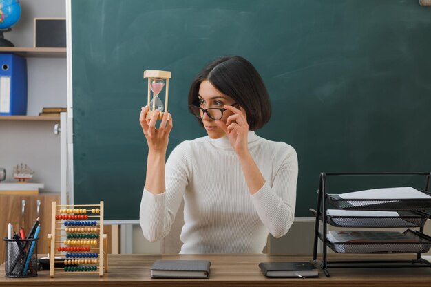 giovane insegnante di pensiero che indossa occhiali che tiene e guarda l'orologio di sabbia seduto alla scrivania con gli strumenti della scuola in classe