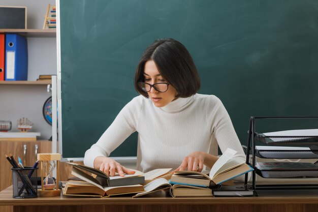 giovane insegnante di pensiero con gli occhiali che legge un libro seduto alla scrivania con gli strumenti della scuola in classe