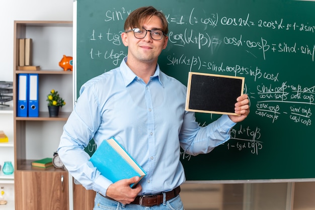 Giovane insegnante maschio che indossa occhiali con libro che tiene piccola lavagna sorridente fiducioso in piedi vicino alla lavagna con formule matematiche in classe