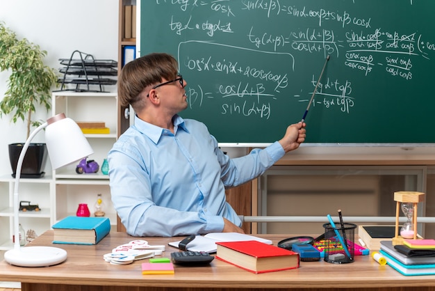 Giovane insegnante maschio con gli occhiali che spiega la lezione guardando fiducioso seduto al banco di scuola con libri e note davanti alla lavagna in classe