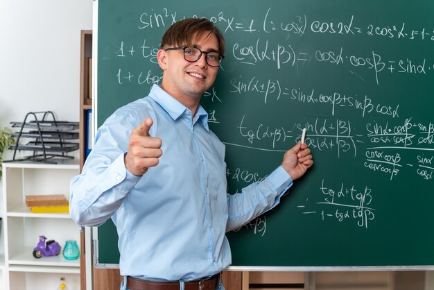 Giovane insegnante maschio con gli occhiali con il gesso che spiega la lezione felice e positiva sorridente in piedi vicino alla lavagna con formule matematiche in classe