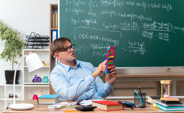 Giovane insegnante maschio con gli occhiali con le fatture che sembra confuso mentre prepara la lezione seduto al banco di scuola con libri e appunti davanti alla lavagna in classe