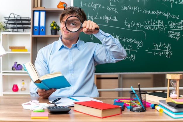 Giovane insegnante maschio con gli occhiali guardando attraverso la lente di ingrandimento seduto al banco di scuola con libri e note davanti alla lavagna in classe