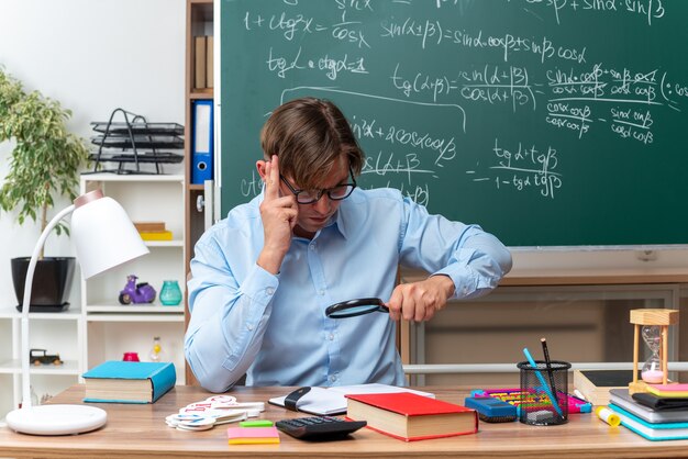 Giovane insegnante maschio con gli occhiali guardando le note attraverso la lente di ingrandimento preparando la lezione seduto al banco di scuola con libri e note davanti alla lavagna in classe