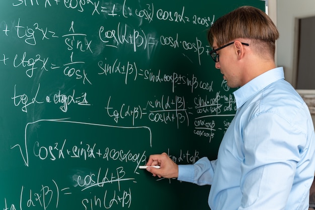Giovane insegnante maschio con gli occhiali in piedi vicino alla lavagna che scrive sulla lavagna formule matematiche che spiegano la lezione guardando fiducioso in classe