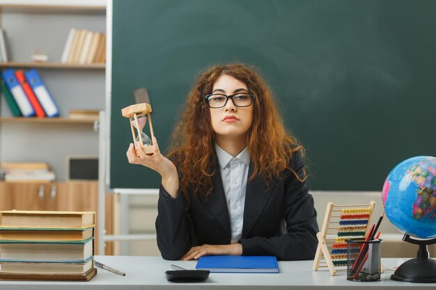 giovane insegnante scontenta che indossa occhiali con orologio di sabbia seduto alla scrivania con strumenti scolastici in classe