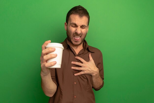 Giovane irritato che tiene la tazza di caffè di plastica che tiene la mano sul petto che mostra la lingua con gli occhi chiusi isolati sulla parete verde