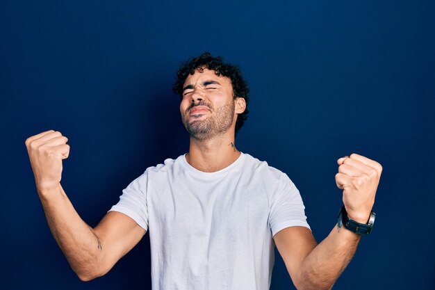 Giovane ispanico che indossa una maglietta bianca casual molto felice ed eccitato facendo il gesto del vincitore con le braccia alzate sorridendo e urlando per il concetto di celebrazione del successo