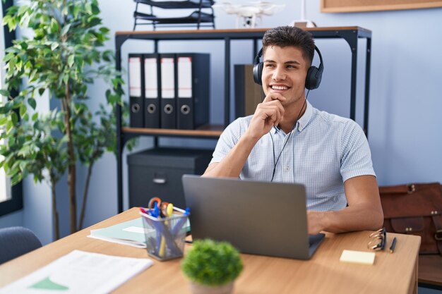 Giovane ispanico che lavora in ufficio indossando le cuffie guardando fiducioso alla telecamera sorridendo con le braccia incrociate e la mano alzata sul mento pensando positivo