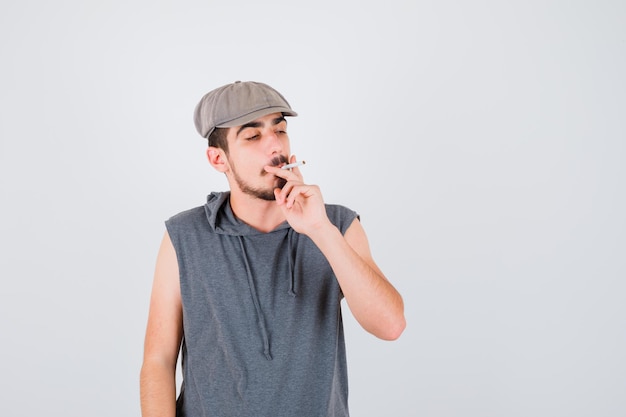 Giovane lavoratore che fuma sigaretta e lo tiene in maglietta grigia e berretto e sembra serio