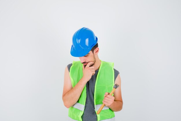 Giovane lavoratore che tiene l'ascia in una mano e sta in posa pensando in uniforme da costruzione e sembra pensieroso