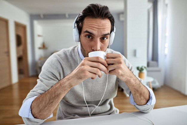 Giovane lavoratore freelance che beve caffè mentre ascolta musica in cuffia a casa