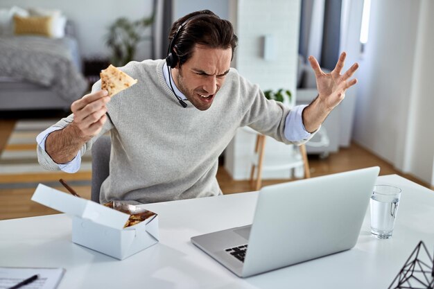 Giovane lavoratore freelance che si sente sconvolto mentre mangia e legge cattive notizie sul laptop a casa