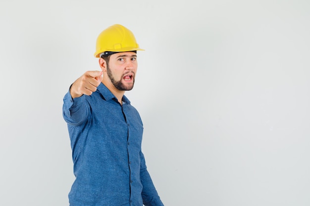 Giovane lavoratore in camicia, casco che punta alla telecamera e che sembra fiducioso