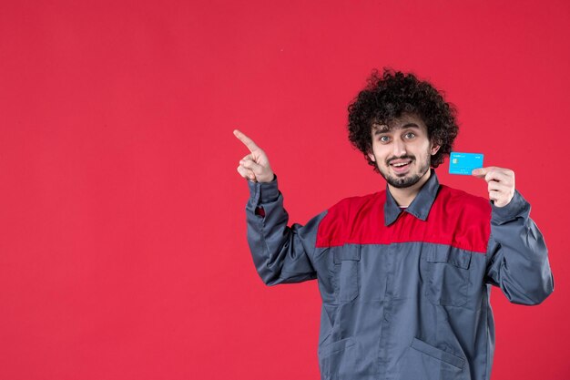 Giovane lavoratore maschio di vista frontale in uniforme che tiene la carta di credito sulla superficie rossa