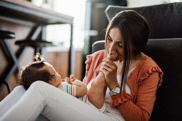 Giovane madre amorevole che si diverte con la sua bambina e bacia i suoi piccoli piedi a casa