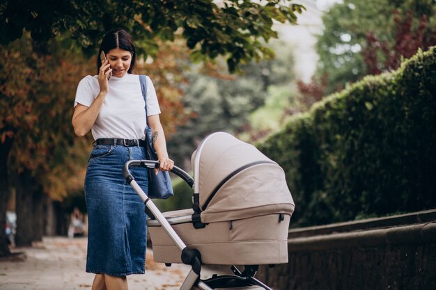 Giovane madre che cammina con il passeggino nel parco e parla al telefono