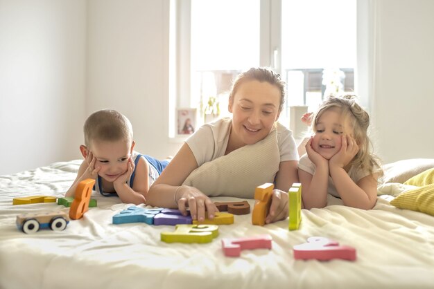 Giovane madre che gioca con i suoi bambini su un letto sotto la luce del sole attraverso le finestre