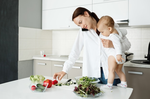 Giovane madre che si prende cura del bambino piccolo, parla al telefono e cucina allo stesso tempo