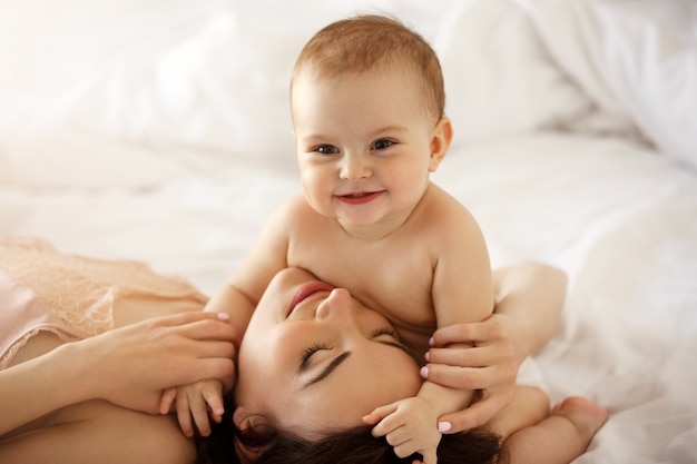 Giovane madre felice e sua figlia del bambino che sorridono abbracciando sdraiato nel letto di casa.
