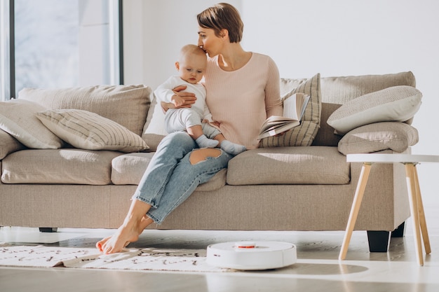 Giovane madre seduta sul divano con il figlio del bambino e guardando l'aspirapolvere robot che fa i lavori domestici