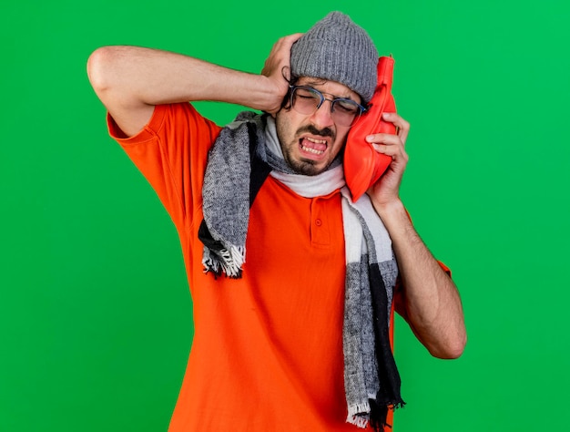 Giovane malato dolorante con gli occhiali cappello invernale e sciarpa che mette la borsa dell'acqua calda sulla testa tenendo la mano sulla testa con gli occhi chiusi isolati sulla parete verde