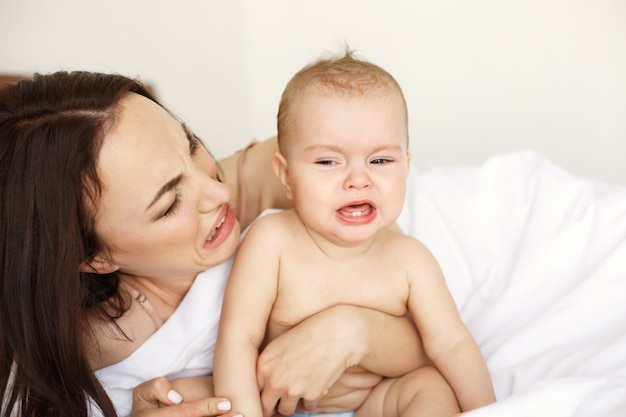 Giovane mamma divertente che gioca la scimmia che il suo bambino piange sdraiato nel letto di casa.