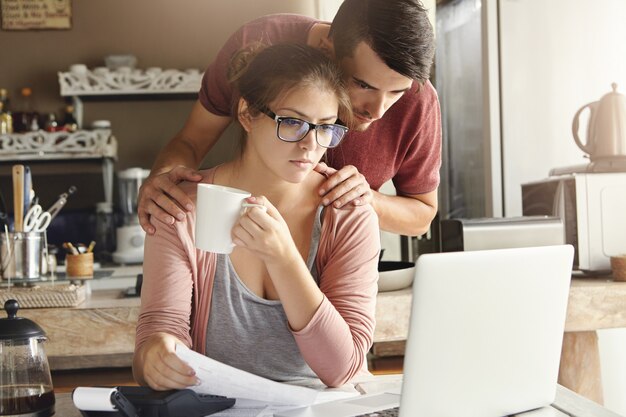 Giovane marito e moglie frustrati che fanno insieme le scartoffie, calcolando le loro spese, gestendo le fatture, usando il computer portatile e la calcolatrice nella cucina moderna