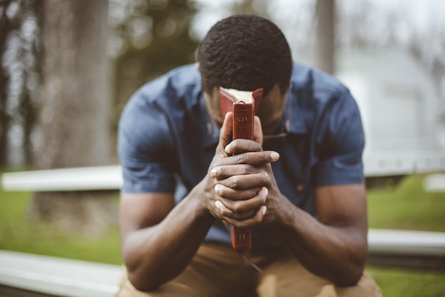 Giovane maschio afro-americano che si siede con gli occhi chiusi con la Bibbia nelle sue mani