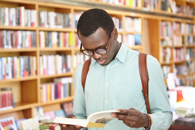 Giovane maschio afroamericano alla moda in camicia e occhiali che guardano attraverso il libro nella condizione della libreria. Turista maschio nero che esplora le librerie locali mentre viaggiando all'estero