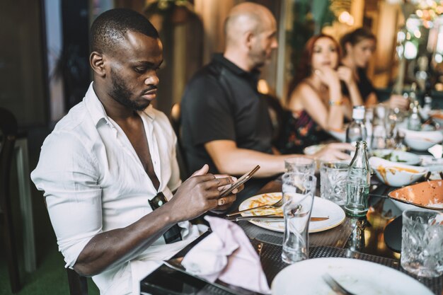 Giovane maschio afroamericano che usa il telefono mentre cena con i suoi amici