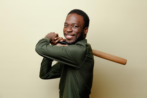 Giovane maschio afroamericano sorridente che indossa la camicia verde che tiene la mazza da baseball isoloated su sfondo bianco