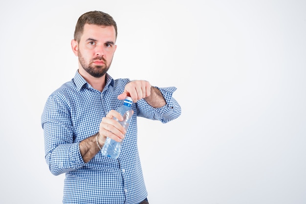 Giovane maschio apertura bottiglia d'acqua in plastica in camicia, jeans e guardando fiducioso, vista frontale.