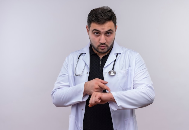 Giovane maschio barbuto medico indossa camice bianco con lo stetoscopio che punta la mano ricordando il tempo preoccupato