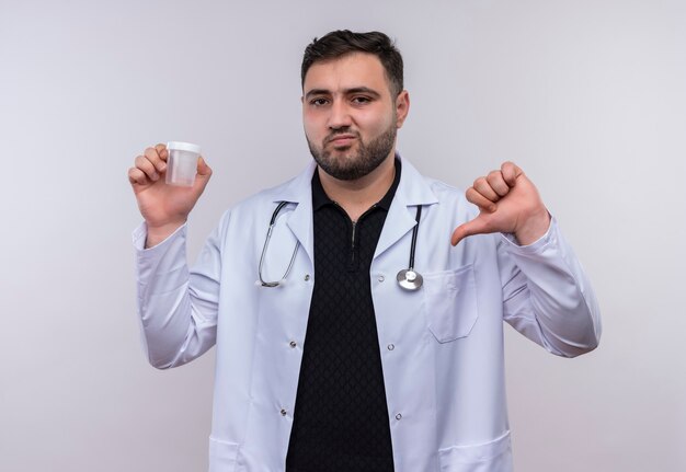 Giovane maschio barbuto medico indossa camice bianco con lo stetoscopio tenendo il barattolo di prova guardando la fotocamera con unhappyface che mostra antipatia