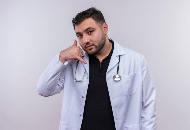 Giovane maschio barbuto medico indossa camice bianco con stetoscopelooking fiducioso facendomi chiamare gesto