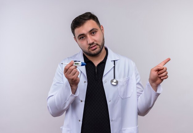 Giovane maschio barbuto medico indossa camice bianco con uno stetoscopio tenendo il termometro che guarda l'obbiettivo con la faccia seria che punta con il dito di lato