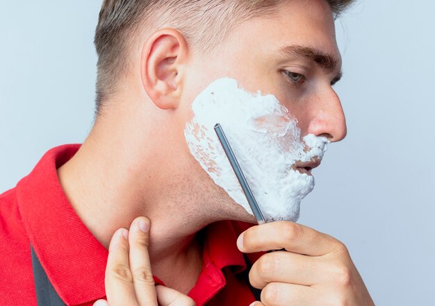 Giovane maschio biondo fiducioso barbiere in uniforme spalmato di fronte con schiuma da barba da barba con il rasoio a mano libera isolato su uno spazio bianco con spazio di copia