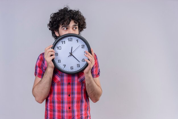 Giovane maschio capelli ricci isolato camicia colorata che si nasconde dietro l'orologio che guarda lontano