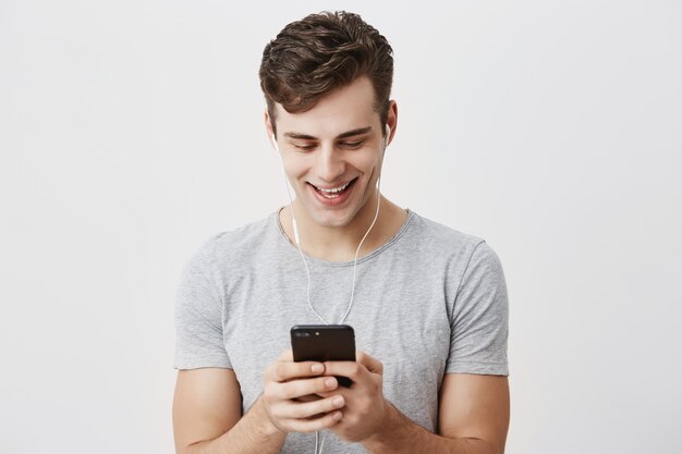 Giovane maschio caucasico bello con capelli scuri e gli occhi azzurri che tengono telefono cellulare, sorridendo con i denti, scrivendo messaggio alla sua amica, indossando le cuffie. Persone, stile di vita e comunicazione moderna.