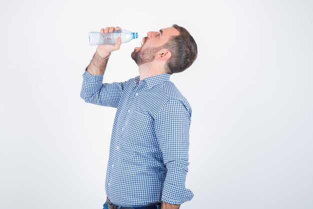 Giovane maschio che beve dalla bottiglia di acqua di plastica in camicia, jeans e che sembra felice, vista frontale.