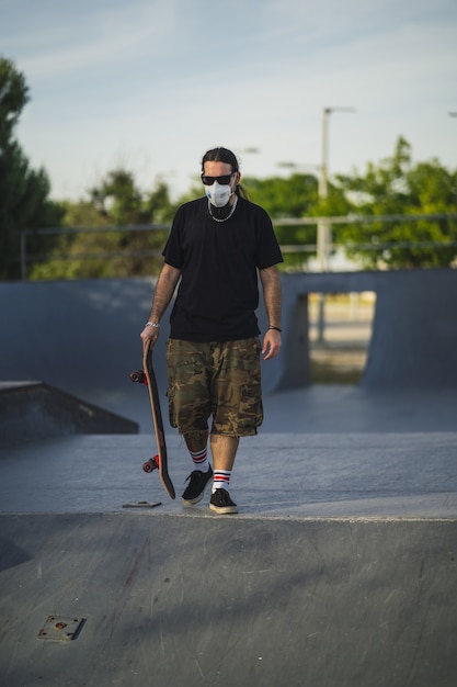 Giovane maschio che cammina in un parco con uno skateboard che indossa una maschera medica - concetto Covid-19