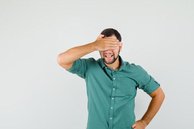 Giovane maschio che copre gli occhi con la mano in camicia verde e sembra timido. vista frontale.