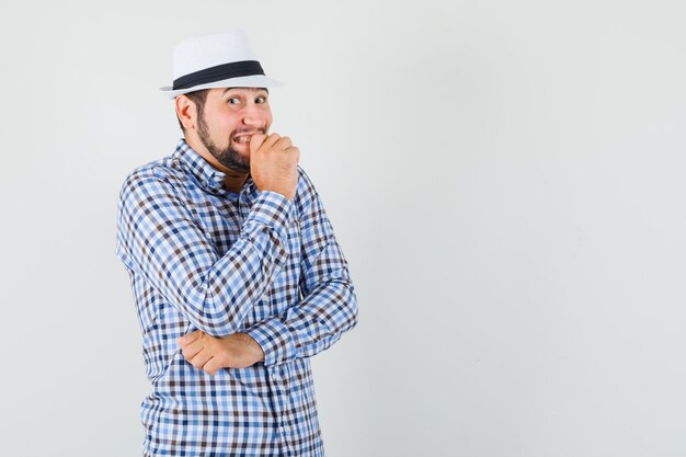 Giovane maschio che guarda l'obbiettivo in camicia controllata, cappello e guardando imbarazzato, vista frontale.