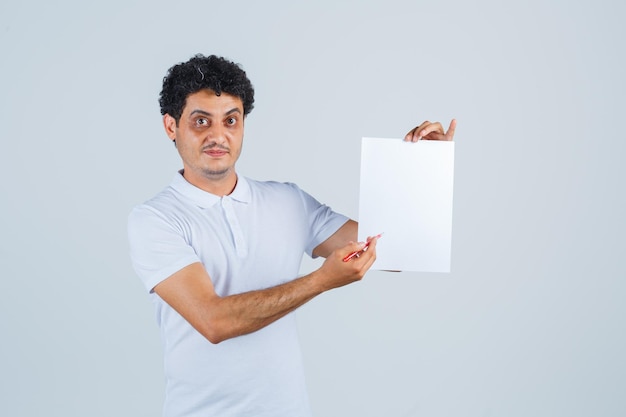 Giovane maschio che indica al foglio di carta con la penna in maglietta bianca e sembra sicuro. vista frontale.