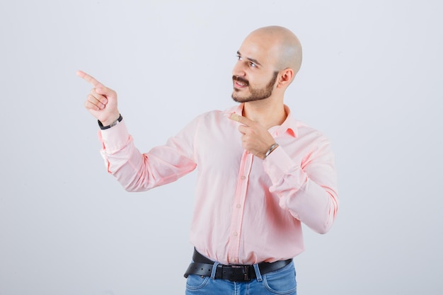 Giovane maschio che indica all'angolo superiore sinistro in camicia, jeans e sembra sicuro. vista frontale.